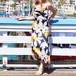 A woman wearing a navy blue maxi dress with a large white and yellow floral print stands on a wooden boardwalk leaning on a white railing, with a beach town background.